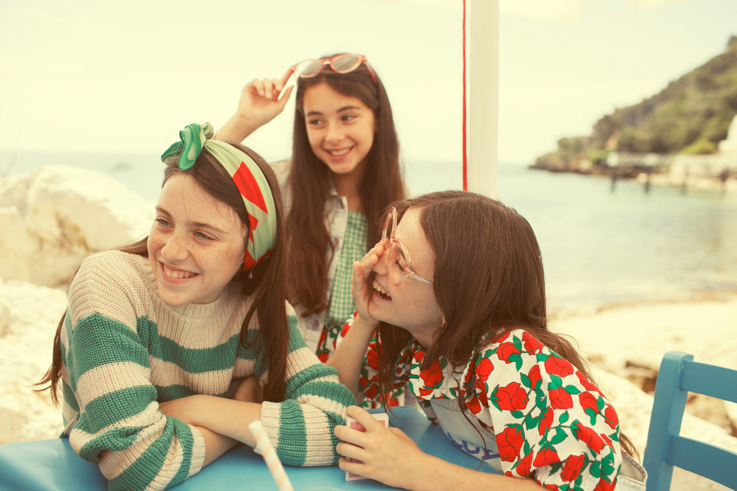 3 niñas vestidas con la colección de verano 2026 de Piupiuchick sentadas en una mesa con la playa de fondo