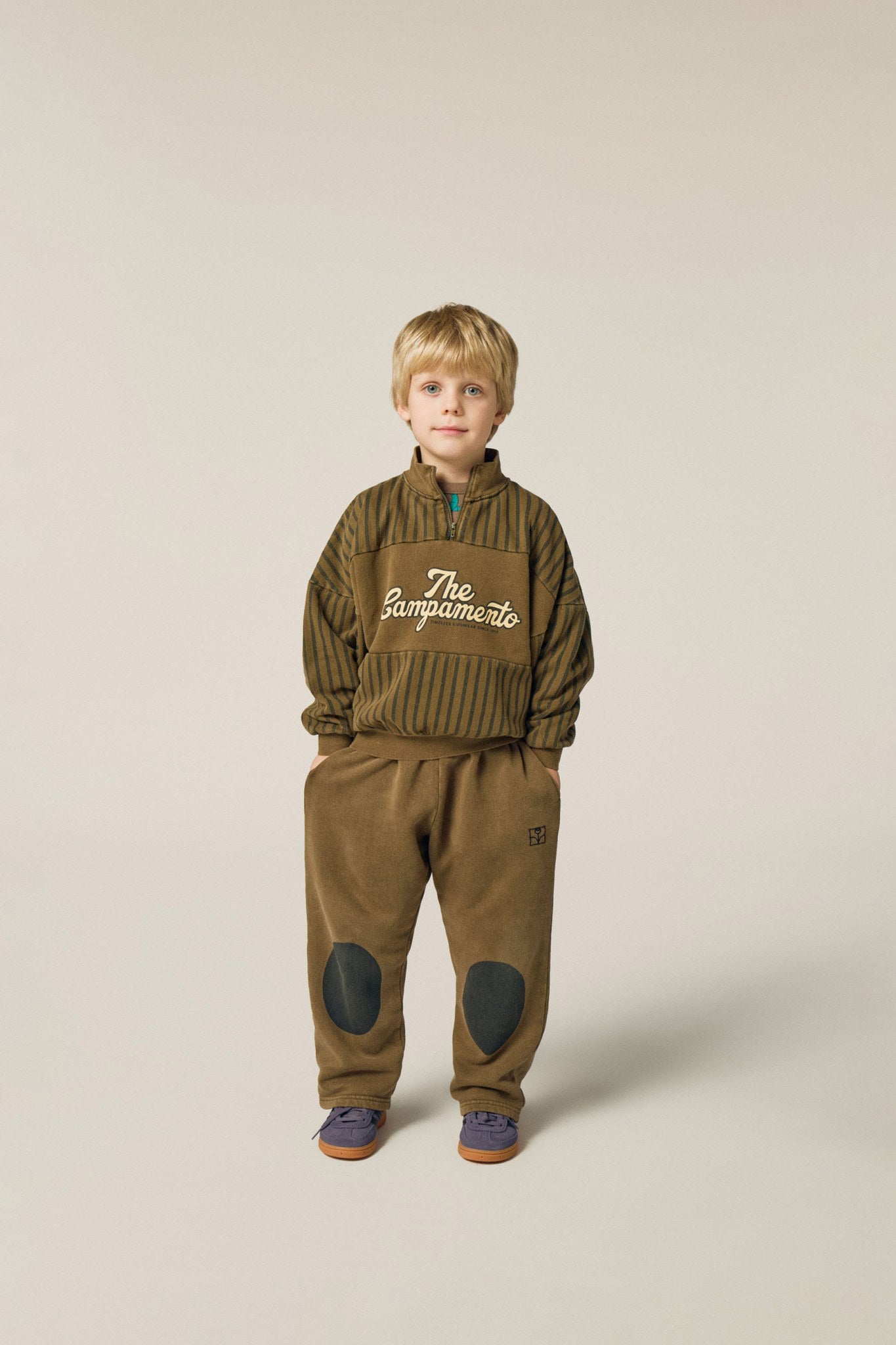 Niño posando de frente con sudadera y pantalón marrón con rodilleras oscuras.
